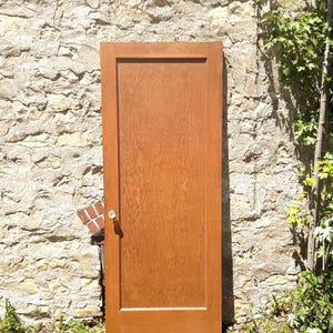 May include: A rectangular wooden door with a medium brown finish and a visible wood grain pattern. The door features a single large central panel and a clear, round glass doorknob attached to the left side of the frame.