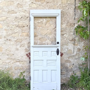 May include: A white, antique door with a rectangular frame and decorative panels. The door features a black doorknob and handle, set against a textured stone wall. The door is weathered, suggesting age and rustic charm.