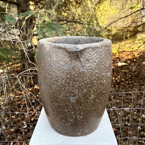 May include: A textured, brown stoneware vessel with a wide opening and a spout-like feature. The antique pot has a cylindrical shape and sits on a white surface. The background features natural greenery and sunlight.
