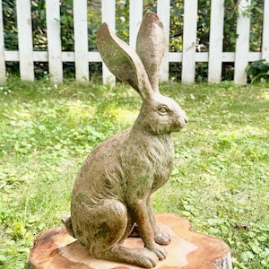 May include: A detailed, bronze-colored rabbit statue with long ears, seated on a tree stump. The sculpture is outdoors, set against a backdrop of green grass and a white picket fence. The rabbit's fur is textured, and the statue has a weathered appearance.