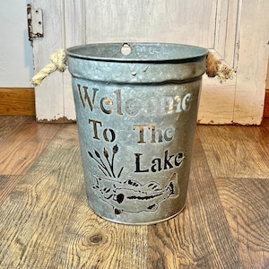 May include: A galvanized metal bucket with rope handles. The bucket features cut-out lettering that reads "Welcome To The Lake" and a fish design. The bucket is sitting on a wooden surface.