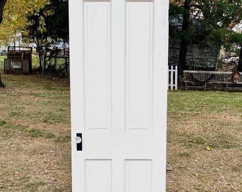 May include: A white four-panel wooden door featuring a rectangular design with two vertical columns of panels. The door includes a black rectangular metal plate surrounding a round glass doorknob positioned on the left side of the door frame.