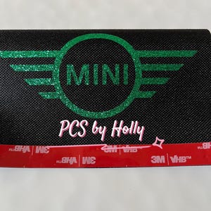 Może przedstawiać: Czarny prostokątny przedmiot z zielonym brokatowym logo MINI i skrzydłami. Tekst "PCS by Holly" jest w różowym skrypcie. Czerwony pasek z tekstem "3M VHB" znajduje się na dole.