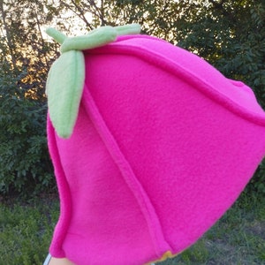 Peut inclure: Un chapeau en polaire rose en forme de fraise avec des feuilles en feutre vert sur le dessus.