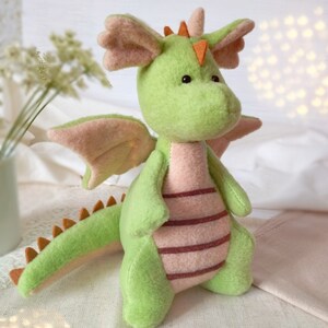 May include: A plush green dragon with pink wings, belly, and ear accents. The dragon has a long tail with orange spikes and is posed on a light-colored surface. The dragon's belly has three horizontal maroon stripes.
