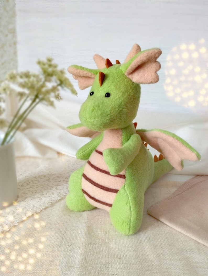 May include: A green plush dragon toy with pink wings and brown stripes. The dragon has black eyes and a small orange horn on its head.