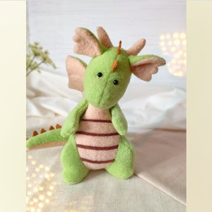 May include: A handmade green dragon plush toy with peach-colored wings, belly, and accents. The dragon has black eyes, orange horns, and brown stripes on its belly. The plush is made of soft fabric and is designed to be a cuddly toy.
