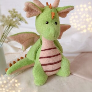 May include: A green plush dragon toy with pink wings and a brown tail. The dragon has a pink and brown striped belly and orange horns.