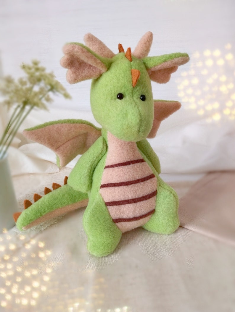 May include: A green plush dragon toy with pink wings and a brown tail. The dragon has a pink and brown striped belly and orange horns.