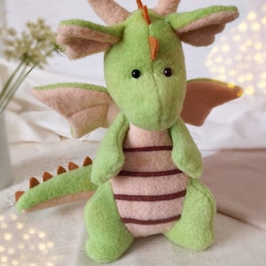 May include: A handmade plush dragon in shades of green and pink. The dragon has large, expressive black eyes, a long tail with orange spikes, and pink wings. The body features brown stripes. The dragon is a soft toy.