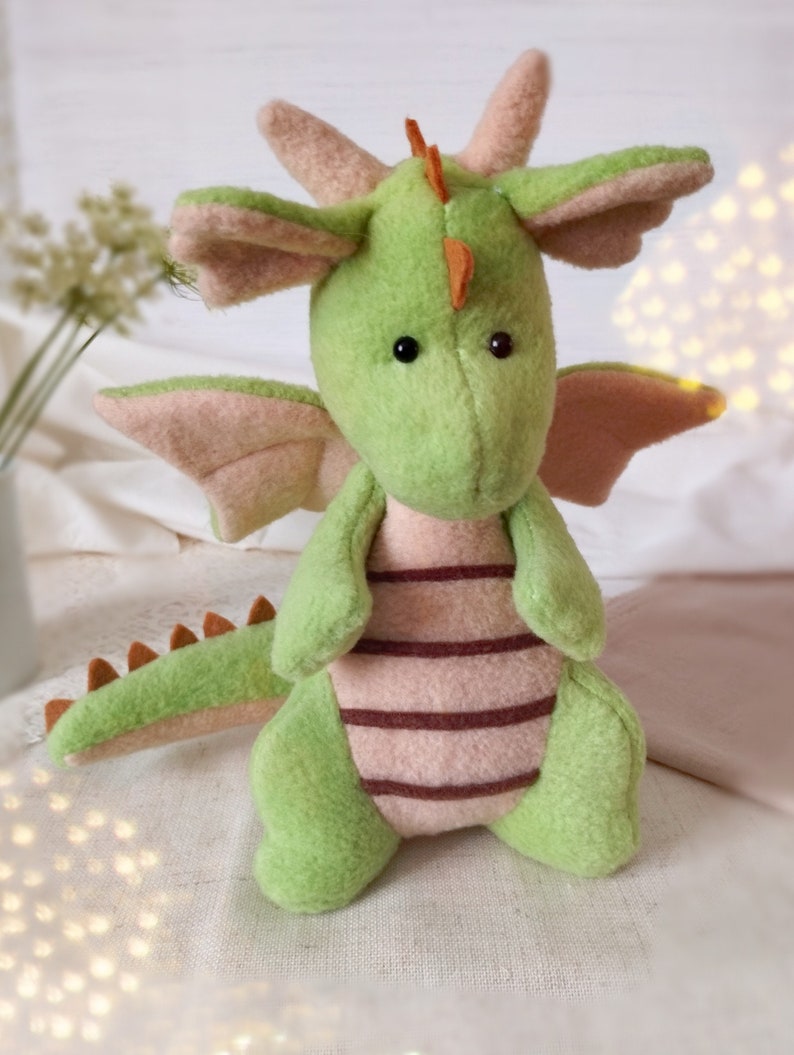 May include: A handmade plush dragon in shades of green and pink. The dragon has large, expressive black eyes, a long tail with orange spikes, and pink wings. The body features brown stripes. The dragon is a soft toy.