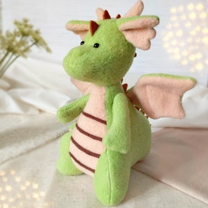 May include: A green plush dragon with pink wings and brown stripes on its back. The dragon has a small, round head with black eyes and a brown nose.