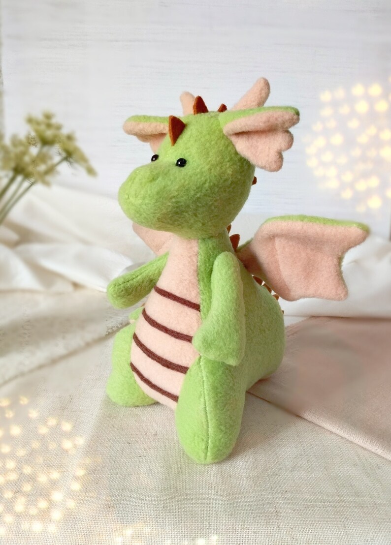 May include: A green plush dragon with pink wings and brown stripes on its back. The dragon has a small, round head with black eyes and a brown nose.
