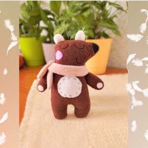 Pode incluir: Um animal de pelúcia marrom com nariz rosa e barriga branca, usando uma echarpe marrom claro. O brinquedo está sentado em uma superfície branca.