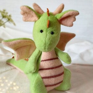 May include: A handmade plush dragon in shades of green and pink. The dragon has large, round eyes, small orange horns, and pink wings and ears. Its belly features horizontal stripes. A whimsical, soft toy.