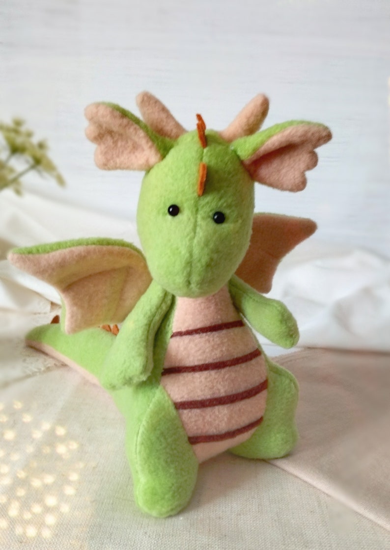 May include: A handmade plush dragon in shades of green and pink. The dragon has large, round eyes, small orange horns, and pink wings and ears. Its belly features horizontal stripes. A whimsical, soft toy.