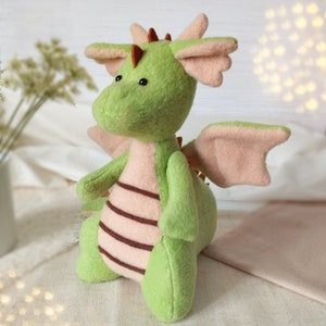 May include: A green plush dragon with pink wings and brown stripes. The dragon has a small, round head with black eyes and a brown nose.