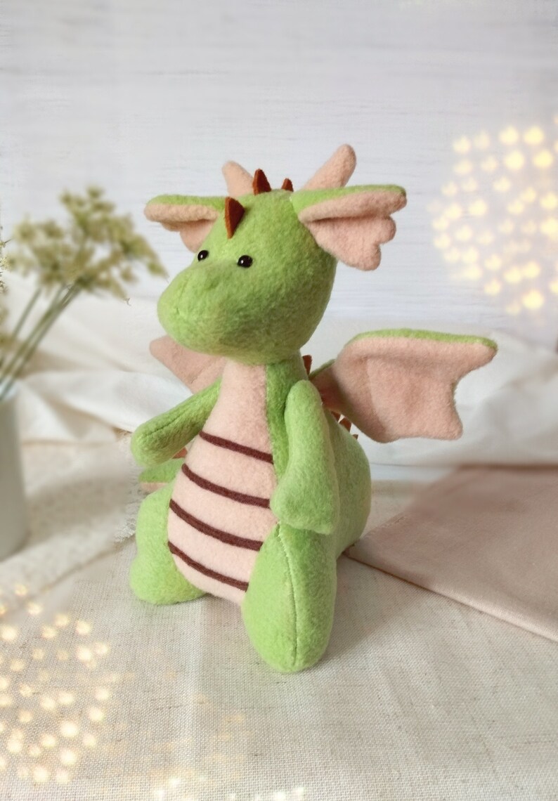 May include: A green plush dragon with pink wings and brown stripes. The dragon has a small, round head with black eyes and a brown nose.