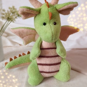 May include: A green plush dragon toy with pink wings and a brown striped belly. The dragon has a small orange horn on its head and black eyes.