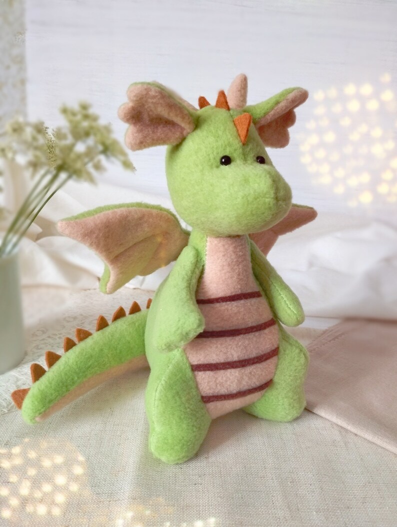 May include: A green plush dragon toy with pink wings and a striped belly. The dragon has orange horns and black eyes.