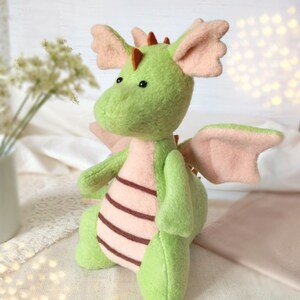 May include: A handmade plush dragon in shades of green and pink. The dragon has large, soft wings, ears, and a belly with brown stripes. It has small black eyes and orange spikes along its back. A whimsical, cuddly toy.