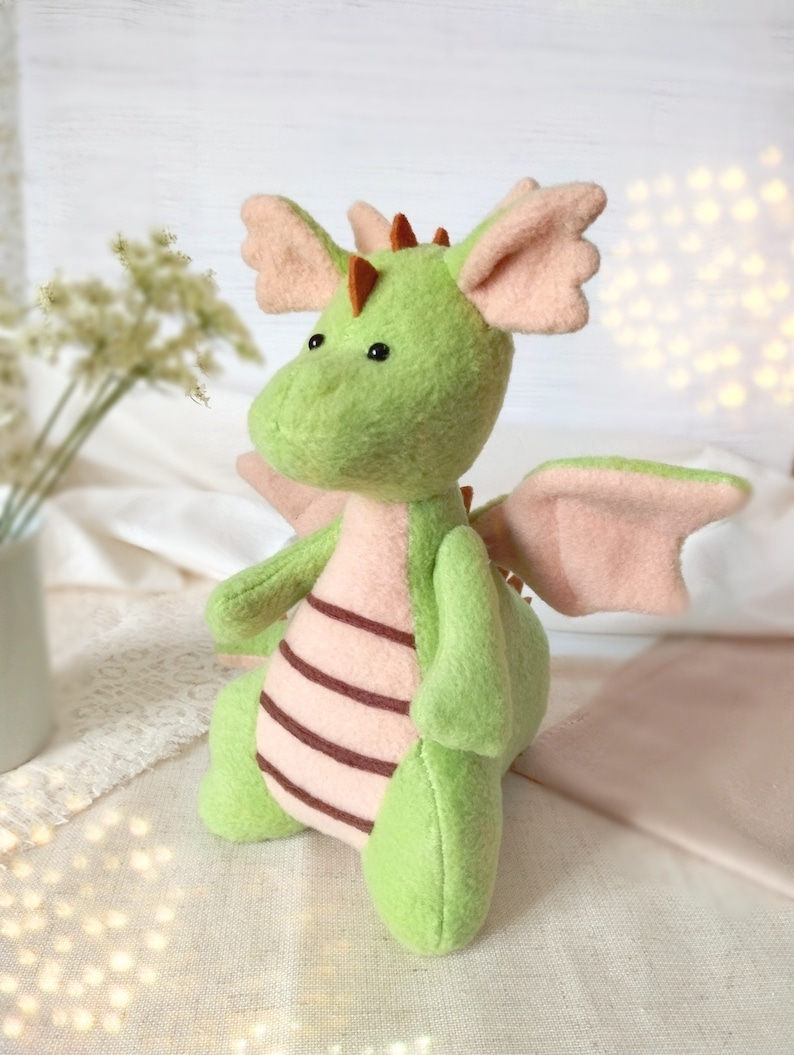 May include: A handmade plush dragon in shades of green and pink. The dragon has large, soft wings, ears, and a belly with brown stripes. It has small black eyes and orange spikes along its back. A whimsical, cuddly toy.