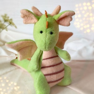 May include: A green plush dragon with pink wings and a striped belly. The dragon has black eyes and an orange horn on its head.