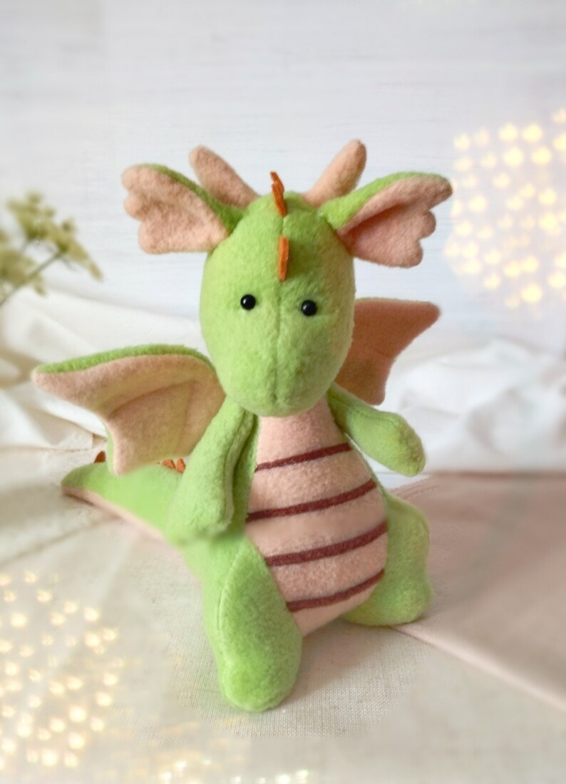 May include: A green plush dragon with pink wings and a striped belly. The dragon has black eyes and an orange horn on its head.