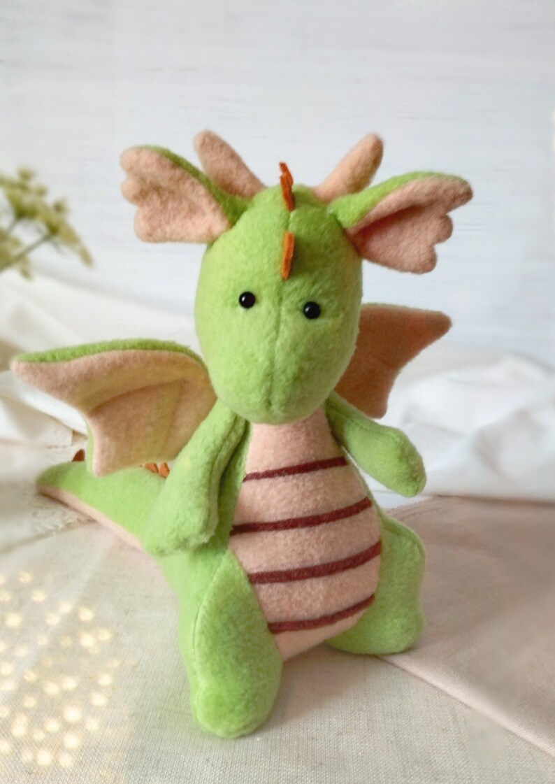 May include: A green plush dragon with pink wings and a striped pink and brown belly. The dragon has black eyes and an orange horn on its head.