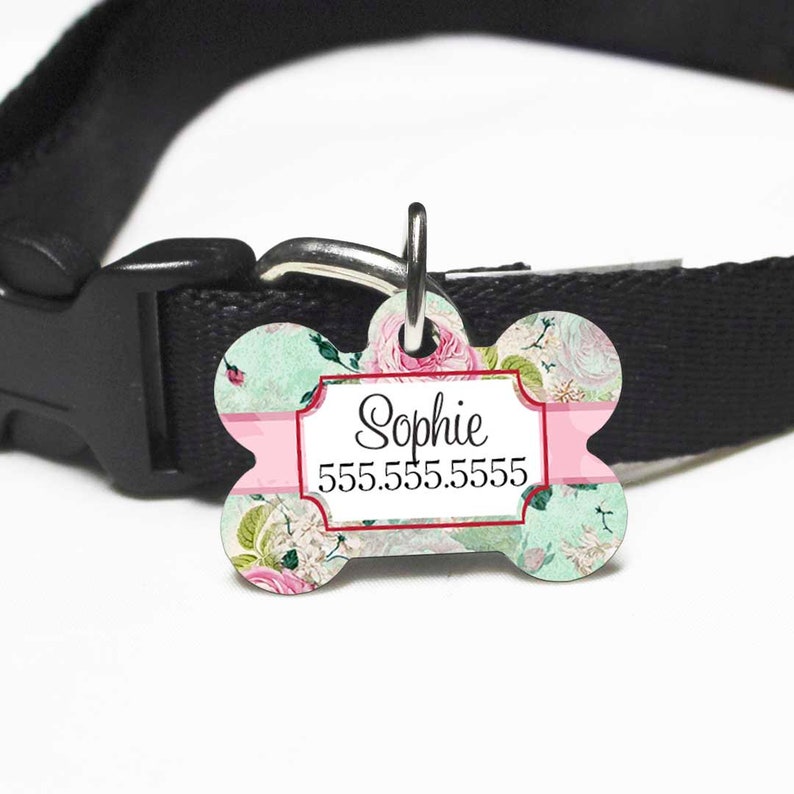 May include: Bone-shaped pet tag with a floral design in shades of pink and mint green. The tag features the name "Sophie" and a phone number in a white rectangular frame. Attached to a black collar.