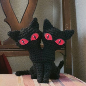 May include: A handmade black crochet cat with two heads and bright red eyes. The cat is sitting on a striped chair. The cat's ears are pointed, and its tail is curled. The cat is a unique, whimsical creation.