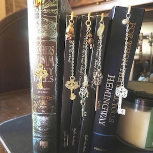 May include: Five metal bookmark charms featuring intricate key designs and dangling beads attached to thin silver and gold chains. Each bookmark is clipped to the top edge of a book spine, including titles like Brothers Grimm Fairy Tales and For Whom the Bell Tolls.