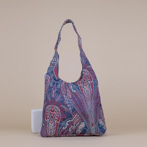 May include: A paisley-patterned shoulder bag in shades of blue, red, and teal. The bag has a single strap and a rounded bottom. The fabric features a detailed, repeating design. The bag is standing upright on a neutral background.