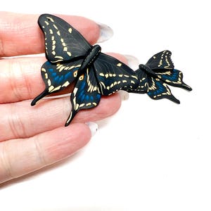May include: Two black butterfly hair clips with detailed wing patterns. The wings display a mix of black, blue, and yellow colours. These clips are designed for hair styling, offering a nature-inspired aesthetic. Each clip is approximately 5 cm in length.