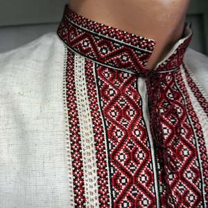 May include: Close-up of a white linen shirt with intricate red, black, and white embroidered details. The shirt features a high collar and decorative vertical panels with geometric patterns. The embroidery adds a traditional touch to the garment.