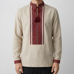 May include: Beige long-sleeve shirt with intricate red and white embroidery on the collar, chest, and cuffs. The shirt features a tassel detail at the neckline and a traditional design. The garment is made of a light-colored fabric.