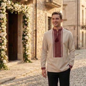 May include: A beige long-sleeve shirt with intricate red embroidery on the collar, chest, and cuffs. The shirt features a tassel detail and is paired with black pants. The setting is an outdoor cobblestone street.