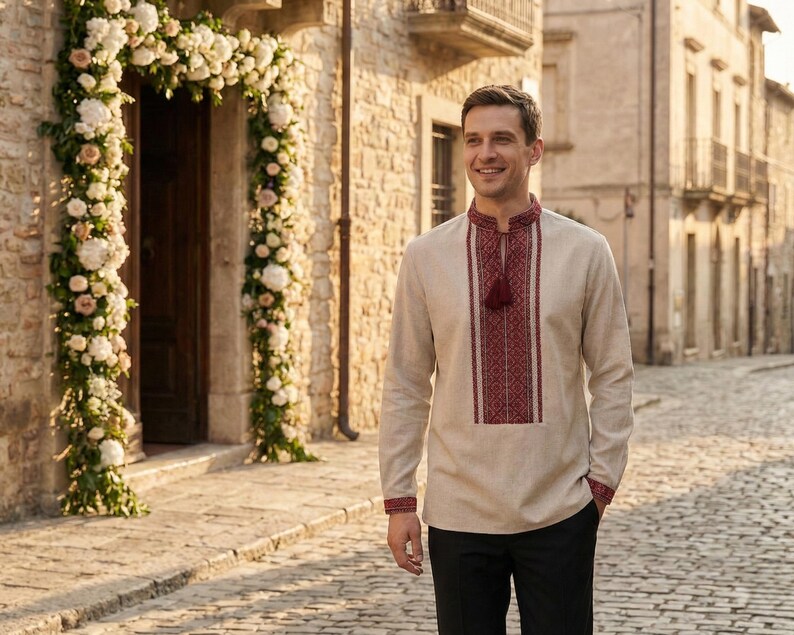 May include: A beige long-sleeve shirt with intricate red embroidery on the collar, chest, and cuffs. The shirt features a tassel detail and is paired with black pants. The setting is an outdoor cobblestone street.