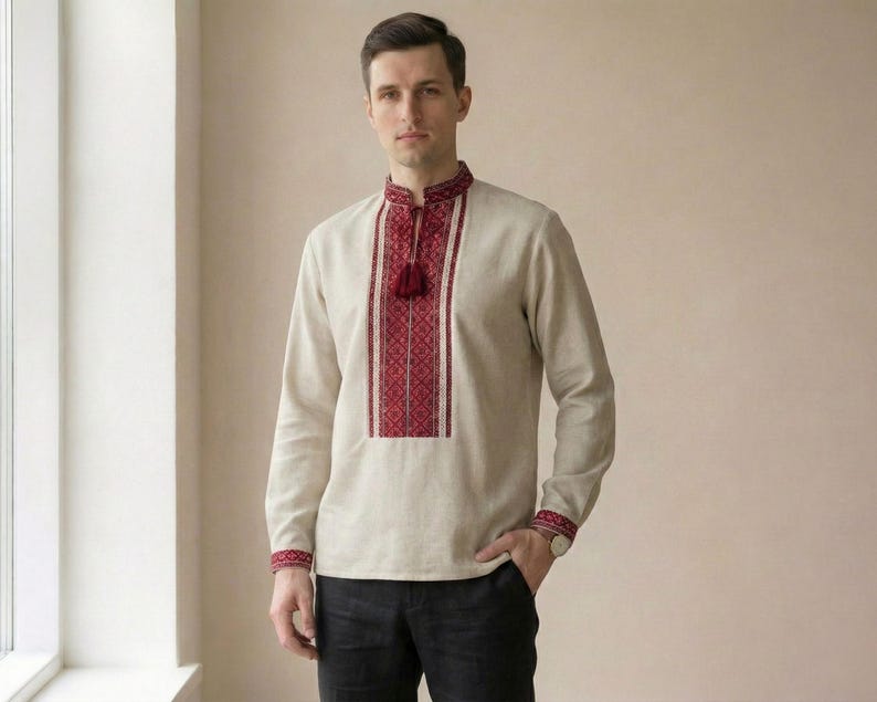 May include: A beige long-sleeve shirt with red and white embroidered details. The shirt features a high collar with a tassel and decorative patterns on the chest and cuffs. The shirt is worn with black pants.