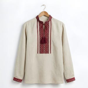 May include: Beige long-sleeved shirt with a mandarin collar and intricate red embroidery. The shirt features a geometric pattern on the chest and cuffs, with a tassel detail at the neckline. It hangs on a wooden hanger against a white background.