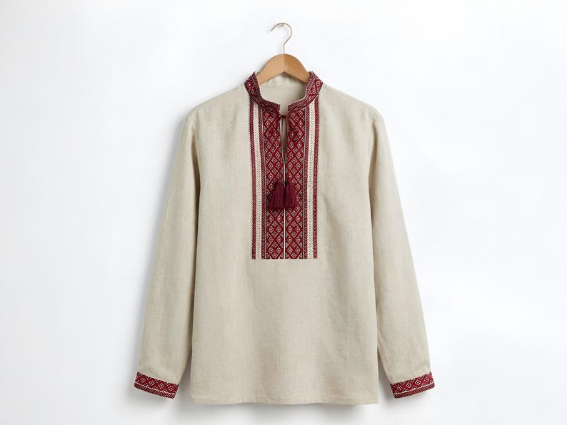 May include: Beige long-sleeved shirt with a mandarin collar and intricate red embroidery. The shirt features a geometric pattern on the chest and cuffs, with a tassel detail at the neckline. It hangs on a wooden hanger against a white background.
