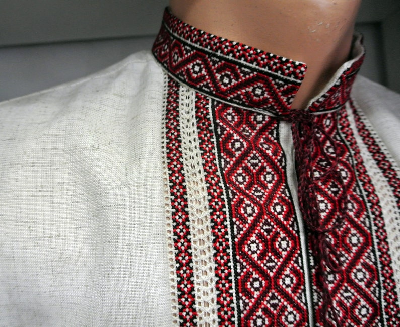May include: Close-up of a white linen shirt with intricate red, black, and white embroidered details. The shirt features a high collar and decorative vertical panels with geometric patterns. The embroidery adds a traditional touch to the garment.