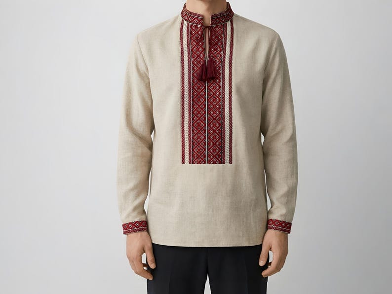 May include: Beige long-sleeve shirt with intricate red and white embroidery on the collar, chest, and cuffs. The shirt features a tassel detail at the neckline and a traditional design. The garment is made of a light-colored fabric.
