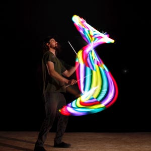 May include: A person is performing with glowing, multi-coloured light trails against a black background. The lights are arranged in a swirling, ribbon-like pattern, with vibrant hues of pink, yellow, green, and blue. The performer is holding two white sticks.