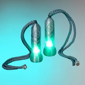 Op de afbeelding: Twee cilindrische poi-handvatten met een getextureerde, zilvergrijze afwerking en geometrische patronen. Elk handvat heeft een felgroen LED-licht dat vanuit de basis schijnt en is bevestigd aan een dik, gevlochten grijs touw met een eindgewicht.