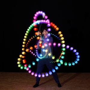 May include: A set of LED poi balls creating long exposure light trails in a circular pattern. The glowing teardrop-shaped lights display a spectrum of colours including purple, pink, blue, green, yellow, and orange against a dark background.