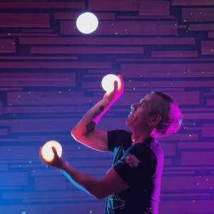 Puede incluir: Una persona hace malabares con tres esferas luminosas. Las esferas son blancas y emiten una luz suave. El fondo es una pared de madera iluminada con tonos morados y azules, creando un ambiente vibrante.