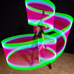 Op de afbeelding: Een persoon is omringd door wervelende lichtsporen in tinten groen, roze en paars. De lichtpatronen creëren een dynamisch, abstract ontwerp. De persoon draagt een donker vest en broek. De achtergrond is zwart.