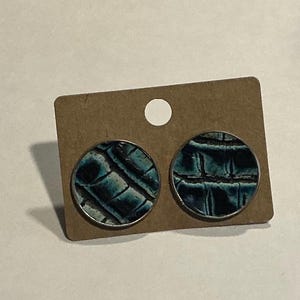 May include: A pair of round stud earrings with a teal and black textured pattern, set on a silver-colored metal. The earrings are displayed on a small, brown cardboard earring card with a white circle at the top.