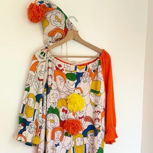 Puede incluir: Un disfraz de payaso vintage con un sombrero a juego, colgado de una percha de madera. El mono blanco está cubierto de caras de dibujos animados y tiene mangas y ribetes naranjas. El sombrero y el mono tienen pompones naranjas.